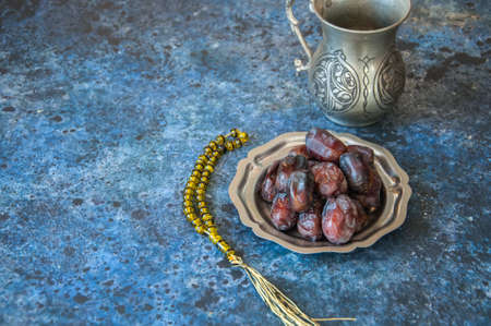 Dried date fruits or kurma served on a old vintage palte with ornaments and beads on a blue stone background, ramadan food concept.の写真素材