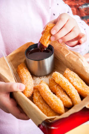 Little girl's hands holding red metal box with Churros sprinkled with sugar and cinnamon and hot chocolate on a craft paper.の写真素材