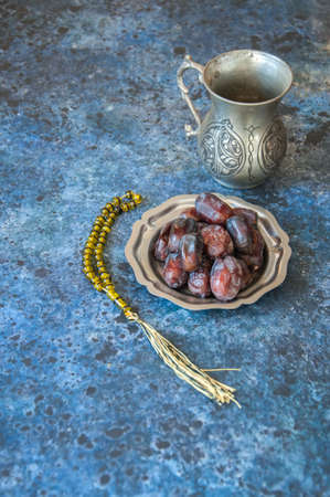 Dried date fruits or kurma served on a old vintage palte with ornaments and beads on a blue stone background, ramadan food concept.の写真素材