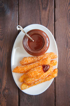 Churros sprinkled with sugar and cinnamon and hot chocolate in a glass jar on a white plate on a wooden background. Traditional spanish treat. Copy Space.の写真素材