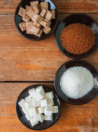 Variety of brown (cane) and white sugar. Granulated and lump sugar in a black small plates on a wooden backgroundの写真素材