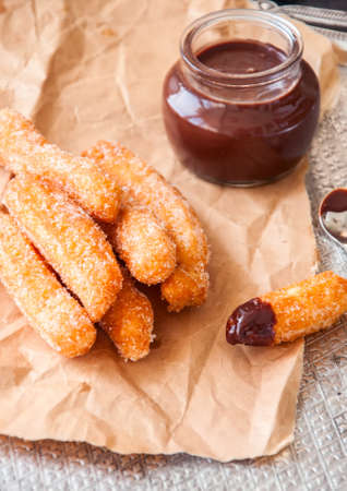Churros sprinkled with sugar and cinnamon on a craft paper and hot chocolate in a glass jar on a metal tray. Traditional spanish treat.の写真素材