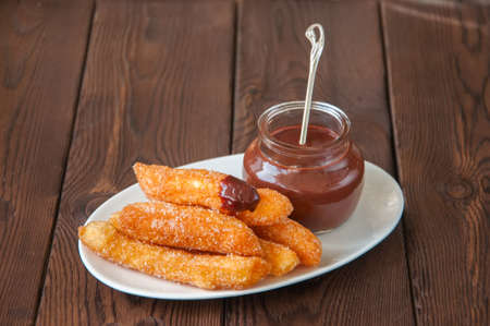 Churros sprinkled with sugar and cinnamon and hot chocolate in a glass jar on a white plate on a wooden background. Traditional spanish treat. Copy Space.の写真素材