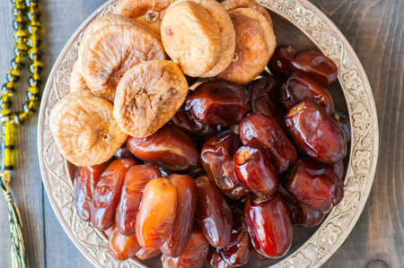 Close up of dried date fruits or kurma and figs served on a old vintage plate with ornaments and beads on a wooden background, ramadan food concept.の写真素材