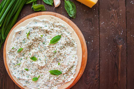 White pizza dough ready to bake on a round wooden board. Different infredients cheese basil leaves garlic on a wooden background. Close up.の写真素材