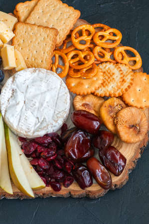 Cheese plate grape salted craker cookies dry dates cranberry, fig, pear pretzels on a wooden board on a black slate background.の写真素材