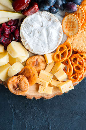 Cheese plate grape salted craker cookies dry dates cranberry, fig, pear pretzels on a wooden board on a black slate background.の写真素材