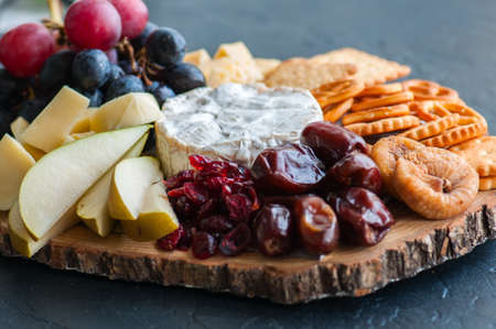 Cheese plate grape salted cracker cookies dry dates cranberry, fig, pear pretzels on a wooden board on a black slate background.の写真素材