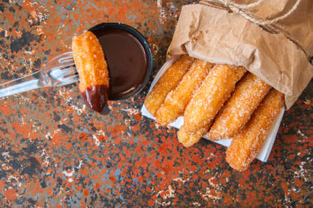 Traditional spanish treat. Popular street food - Churros sprinkled with sugar and cinnamon served in a craft paper bag with hot chocolate sauce in can tin on rusty background. Copy space.の写真素材