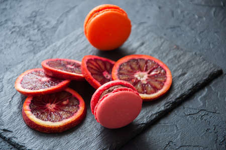 Whole and slices of blood oranges and macarons with chocolate ganache filling on a black slate background. Copy space and close up.の写真素材