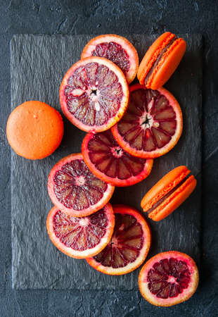 Whole and slices of blood oranges and macarons with chocolate ganache filling on a black slate background.Top view and close up.の写真素材