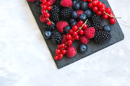 Berry raspberries red currants and blueberries on black slate board over white stone background. ...