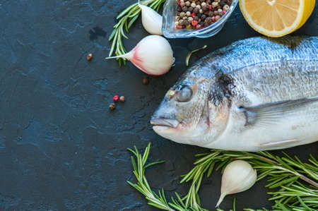 Fresh raw dorado fish with rosemary, garlic and lemon and pepper on a black slate background. Selective focus. Overhead view. Copy space.の写真素材