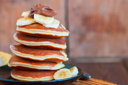 Close up of stack of homemade pancakes with bananas and chocolate paste in a plate on a board....