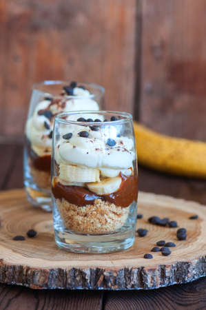 Dessert in a glass - Banoffee pie from bananas, crumble, salted caramel and wipped double creamand chocolate drops on a board. Wooden background, close up and copy space.の写真素材