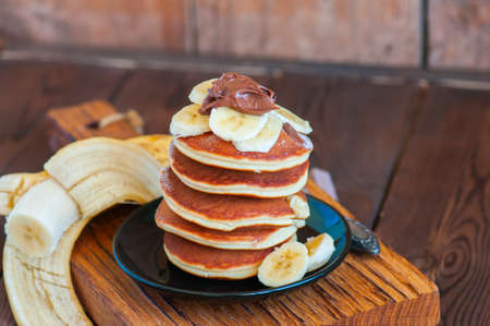 Close up of stack of homemade pancakes with bananas and chocolate paste in a plate on a board. Wooden background. Healthy breakfast and food concept. の写真素材