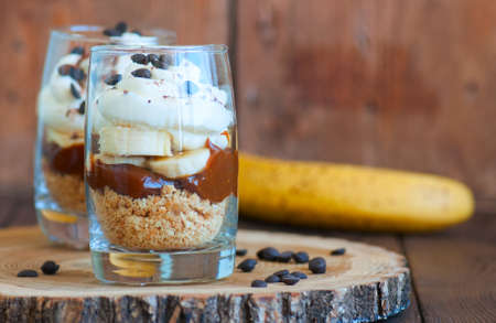 Dessert in a glass - Banoffee pie from bananas, crumble, salted caramel and wipped double creamand chocolate drops on a board. Wooden background, close up and copy space.の写真素材