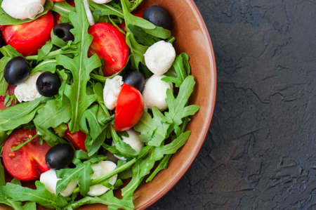 Close up of fresh salad with rucola, tomatoes cherry, olives, spring onion, mozzarella and spices in a bowl on a black slate background with copy space. Healthy food, Diet, Detox, Clean Eating or Vegetarian conceptの写真素材