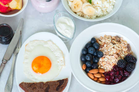 Healthy breakfast concept. Homemade granola (muesli) with blackberry, dry cranberry, blueberry, almond, cottage cheese with banana, fried eggs with bread, yogurt in a jar, sour cream on a white background.の写真素材