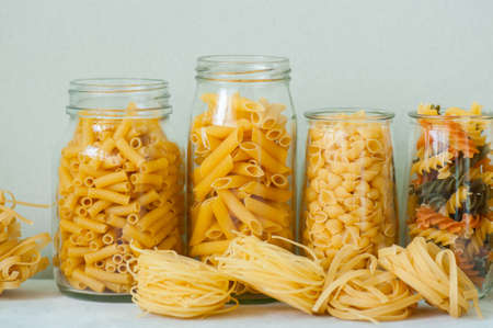 Assortment of different shapes whole grain raw Italian pasta in a glass jar. White stone background. Copy space. Cooking concept.の写真素材