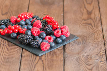 Mix of berries raspberries red currants blueberries and blackberries on black slate board. Wooden...