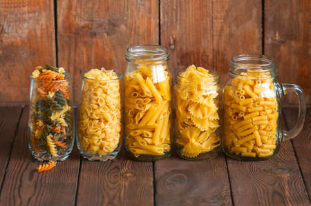 Assortment of different shapes whole grain raw Italian pasta in a glass jar. Wooden background. Copy space. Cooking concept.の写真素材