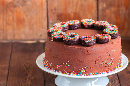 Close up of homemade rich chocolate cake with chocolate ganache frosting decorated with baked doughnuts with confetti glaze on a white cake stand on a wooden background. Copy space.の写真素材