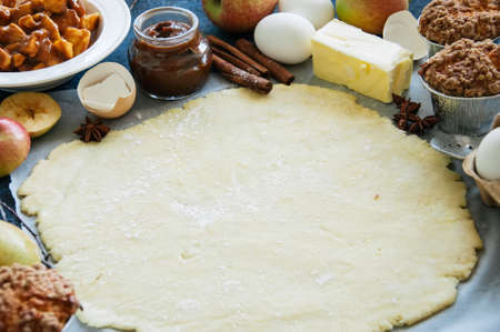 Set of ingredients for baking, raw dough for pie, spices, apple pie filling, caramel, eggs and butter on a blue table. Top view and copy space.の写真素材
