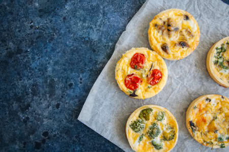Set of savory mini tarts on a baking paper. Vegetable quiches with tomatoes, mushrooms, herbs, broccoly. Blue stone background. Close up and copy space.の写真素材