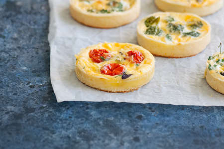 Set of savory mini tarts on a baking paper. Vegetable quiches with tomatoes, mushrooms, herbs, broccoly. Blue stone background. Close up and copy space.の写真素材