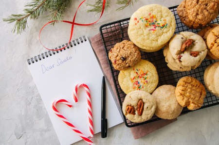 Stack of different cookies on a wire rack. Candy canes and white page of notepad with inscription...