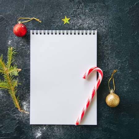 Empty white page of notepad, fir tree branch, candy cane, balls on a black snowy background. Flat...