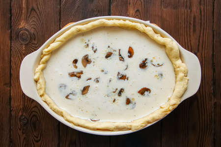 Half-finished quiche with mushrooms and cheese ready to bake in a ceramic dish on a wooden background.の写真素材