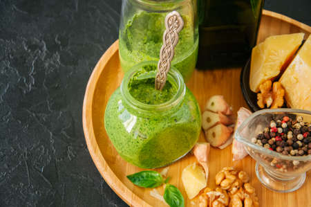 Basil walnut pesto in a jar with raw ingredients on a wooden plate. Black stone background. の写真素材