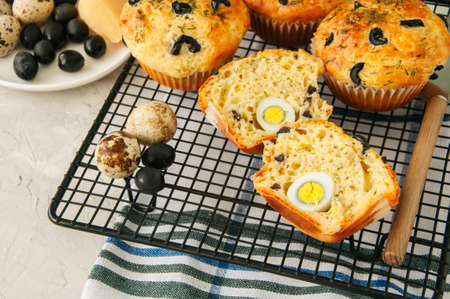 Savory muffins with olives, quail eggs and herbs on a wire rack on a white stone backdrop. Rustic style.の写真素材