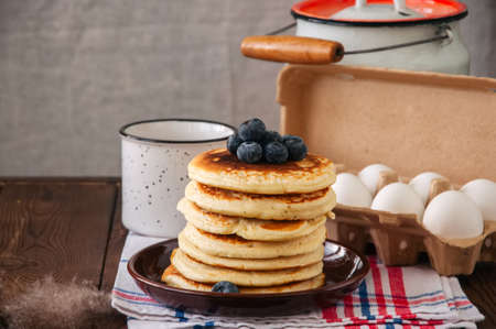 Stack of homemade pancakes with blueberries in a plate, milk in a flask, eggs in a paper container. Rustic style, still life. Copy space.の写真素材