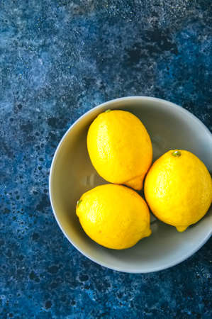 Three ripe yellow lemons in a grey bowl on a blue stone nackground. Top view and copy space.の写真素材
