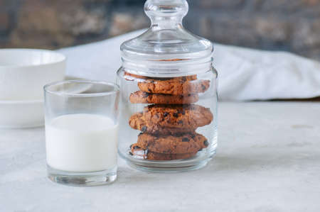 Homemade chocolate chip cookies in a jar and glass of fresh milk on a white stone background.