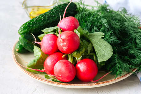 Fresh young vegetables for healthy salad. Radishes cucumbers dill spices and oil on a plate. White stone background. の写真素材