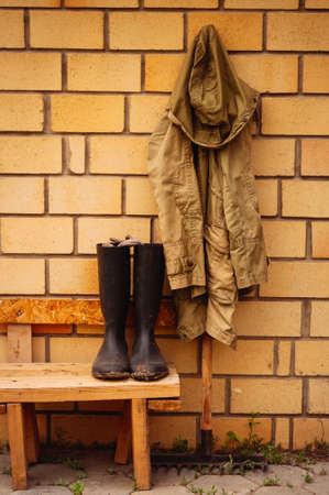 Farmer kit - rain coat haning on rake and rubber boots on a bench. Toned.