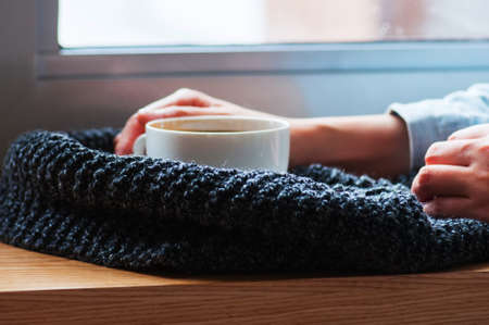 Woman's hand holding americano coffee on woolen scarf on windowsill.