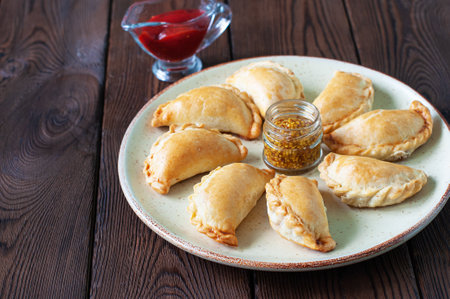 Freshly baked small empanadas on a plate with ketchup and mustard on a wooden background.の写真素材