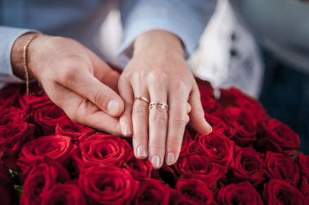 guy holds the girl's hand with a wedding ring over a bouquet of red rosesの写真素材