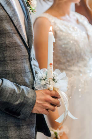 The bride and groom hold shining candles during the ceremony in the church. Hands of newlyweds with...