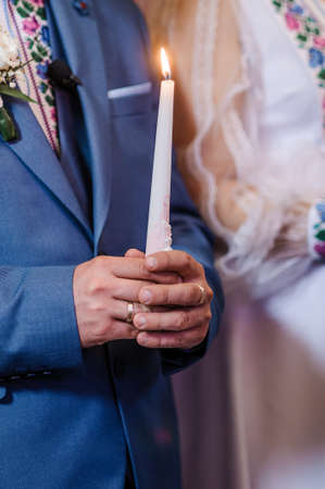 The bride and groom hold shining candles during the ceremony in the church. Hands of newlyweds with candles in the church. Church religious details. Traditionsの写真素材