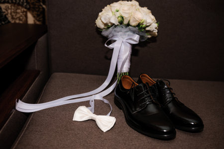 White bow tie, black classic shoes and bridal bouquet with white roses on a brown background