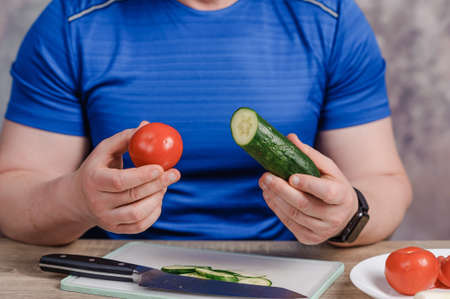 A man holds a cucumber and a tomato in his hands. There is a knife on the board, and a cut cucumber on the tableの写真素材