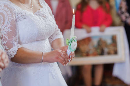The bride and groom hold shining candles during the ceremony in the church. Hands of newlyweds with candles in the church. Church religious details. Traditionsの写真素材
