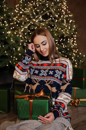 A girl in a sweater with a deer sits on the floor and holds a green gift box. Christmas trees in the backgroundの写真素材