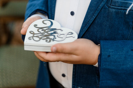 A pair of gold wedding rings in a white wooden box in the shape of a heart in the hands of a guy.